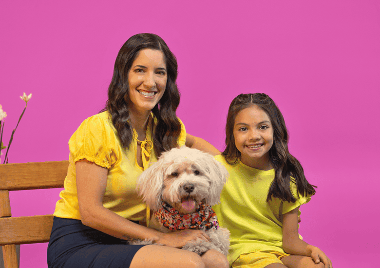 Mujer y niña vestidas de amarillo sentada en banca con perro blanco en el regazo con fondo rosa