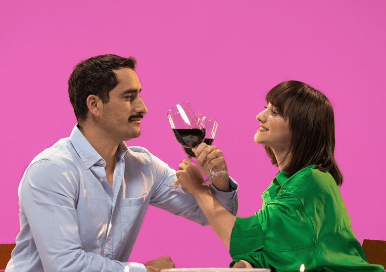 Pareja de hombre con camisa azul y mujer con camisa verde brindando con vino teniendo brazos cruzados en un fondo rosa