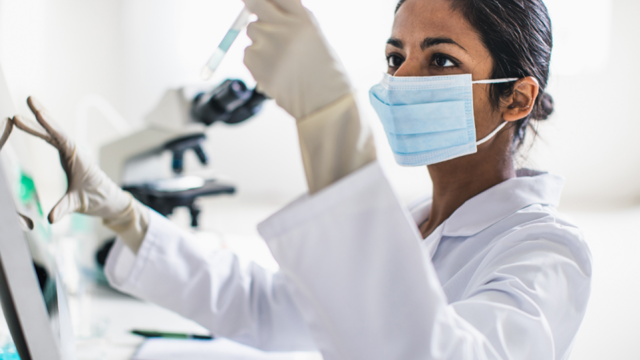 woman-looking-at-beaker Scientist in a mask and lab coat looking at beaker