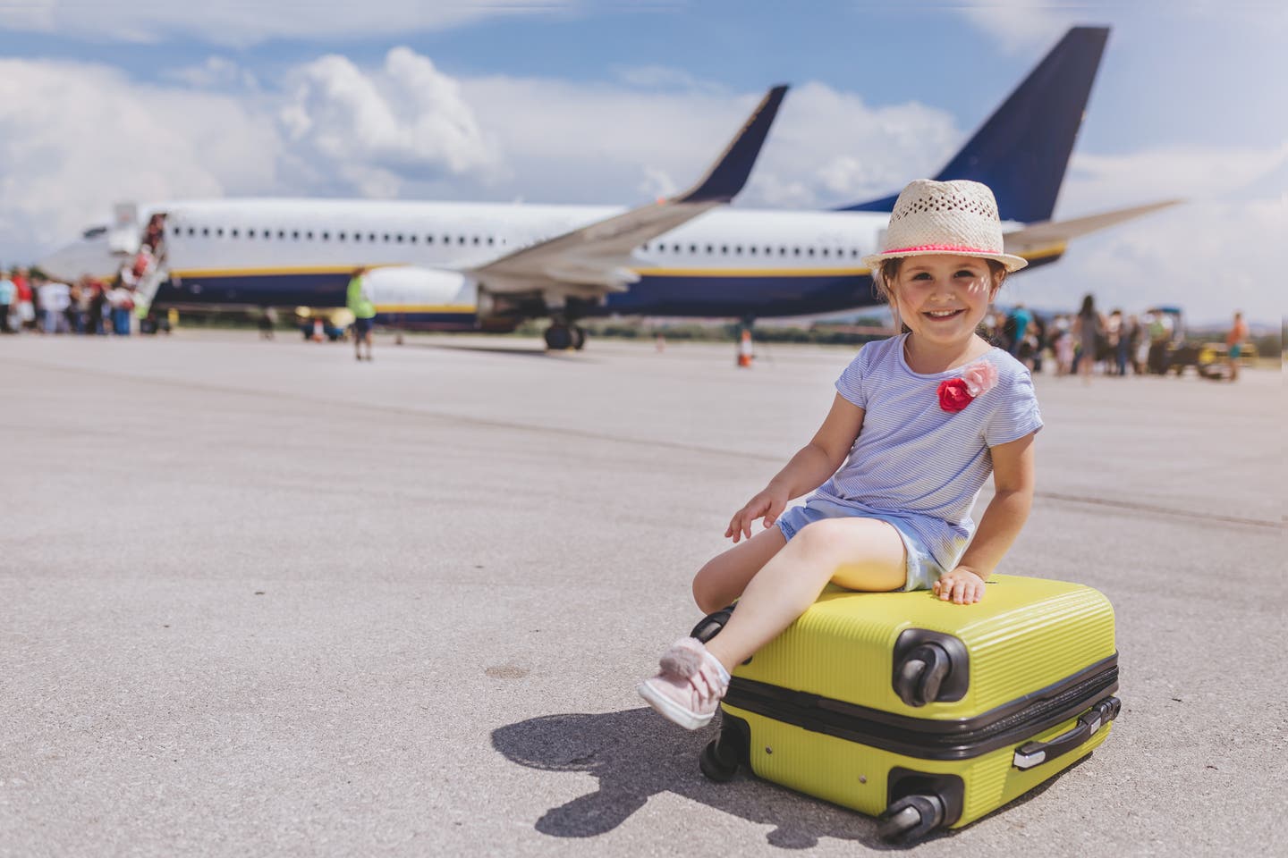 Mädchen sitzt auf einem gelben Koffer vor einem Flugzeug