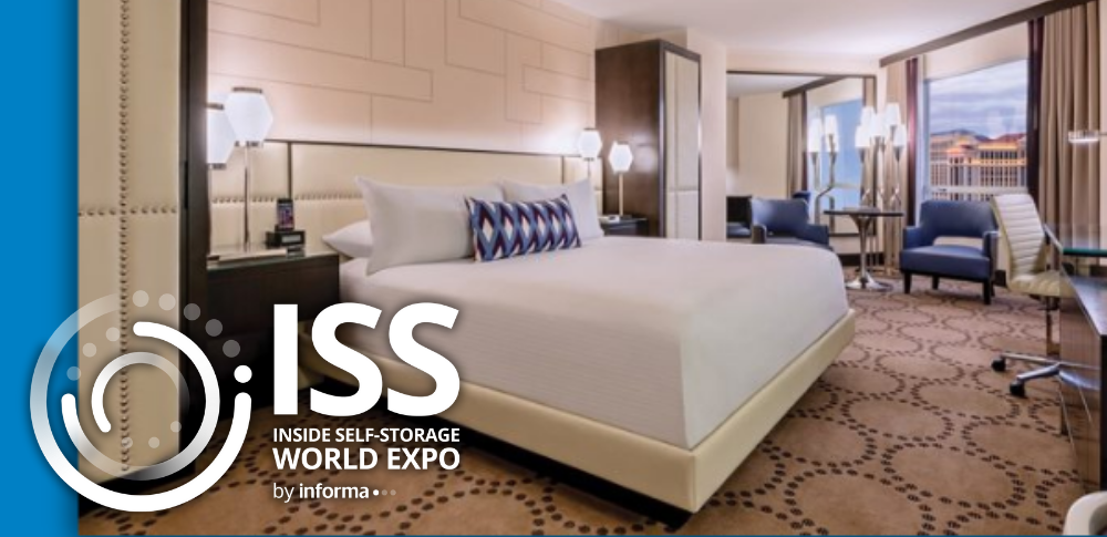 Modern hotel room with a large bed, geometric carpet, stylish lighting, blue chairs by the window, and cityscape view.