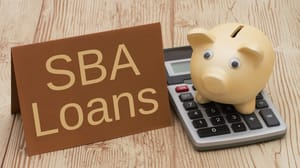 A sign that says SBA Loans on a desktop next to a piggy bank and calculator A sign that says SBA Loans on a desktop next to a piggy bank and calculator