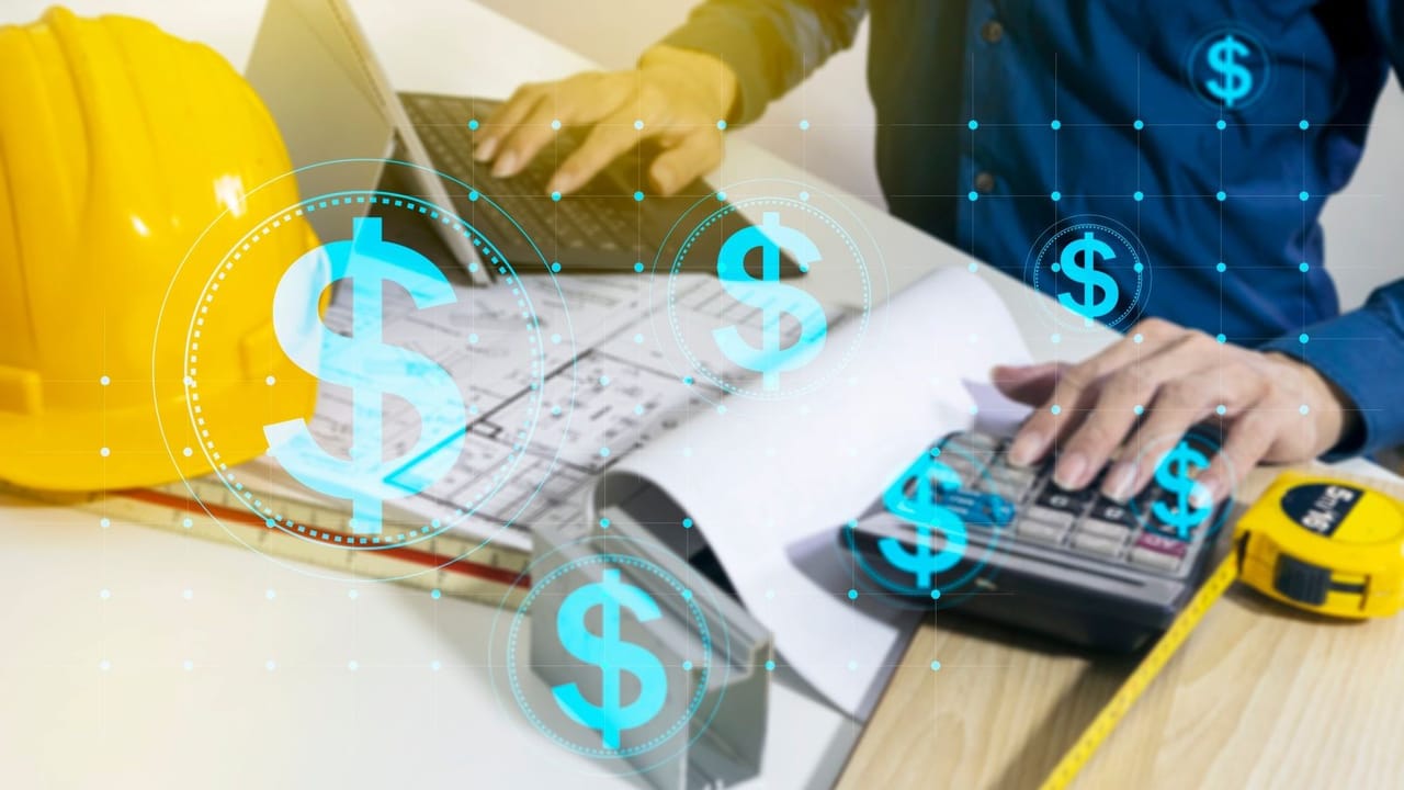 A person working on a laptop and calculator on a desk with design plans and construction materials next to them A person working on a laptop and calculator on a desk with design plans and construction materials next to them