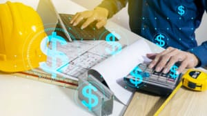 A person working on a laptop and calculator on a desk with design plans and construction materials next to them A person working on a laptop and calculator on a desk with design plans and construction materials next to them