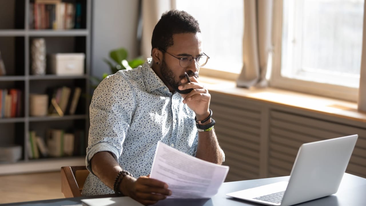 An image of a man looking at a computer screen while holding paper with information An image of a man looking at a computer screen while holding paper with information