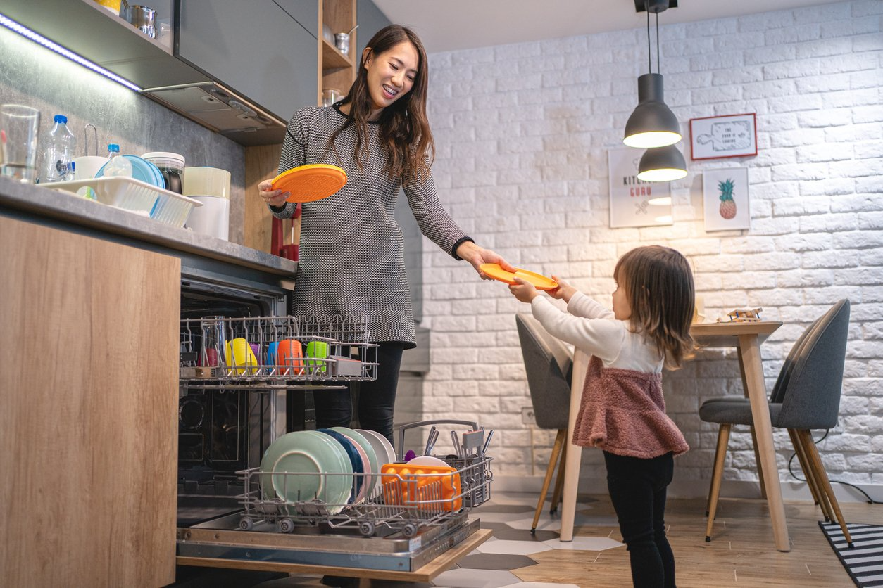 A woman and a child unloading a dishwasher in a cozy kitchen. The woman smiles as she hands a yellow plate to the child.
