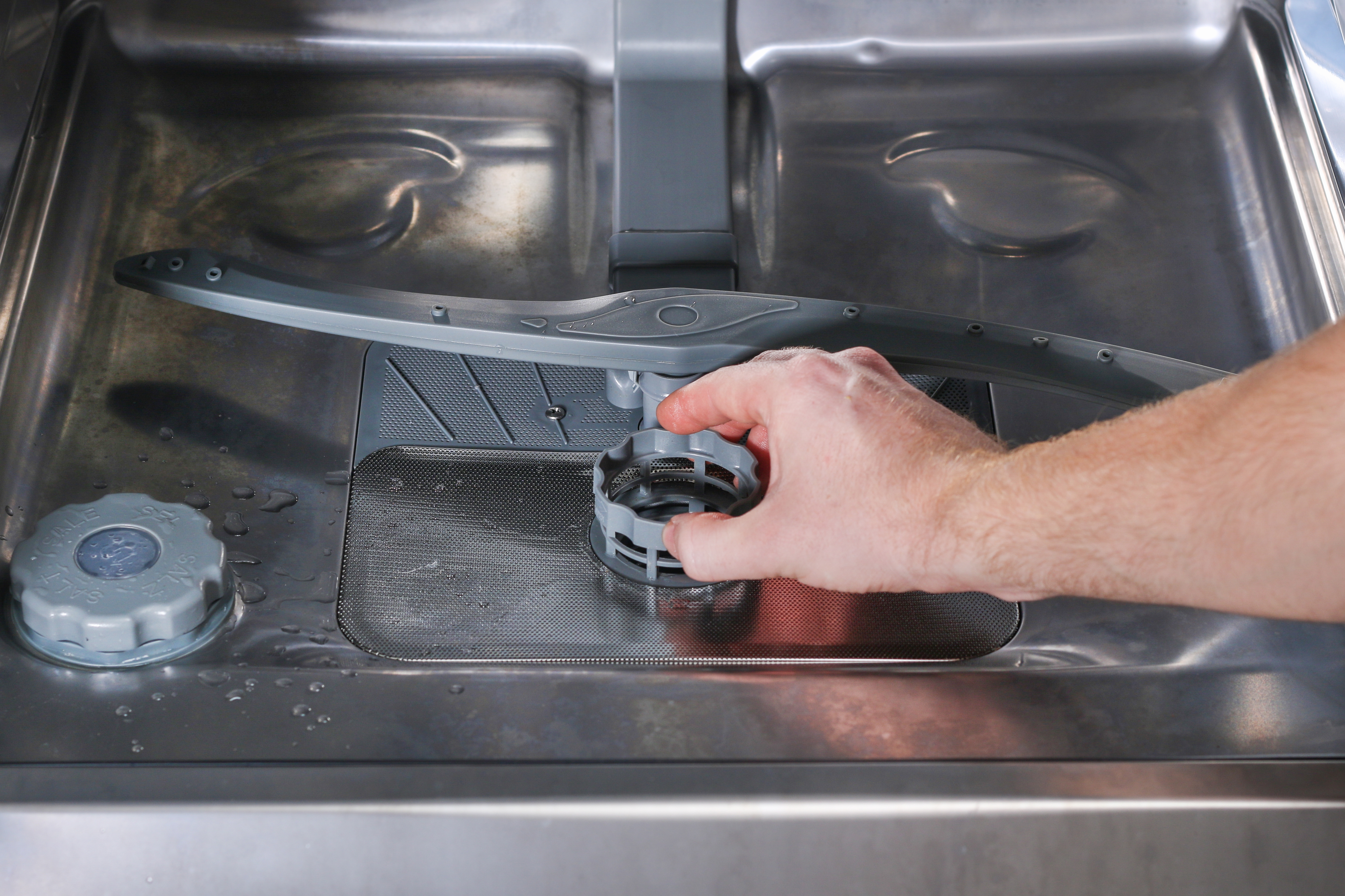 A hand unscrews the dishwasher filter at the base of the machine.