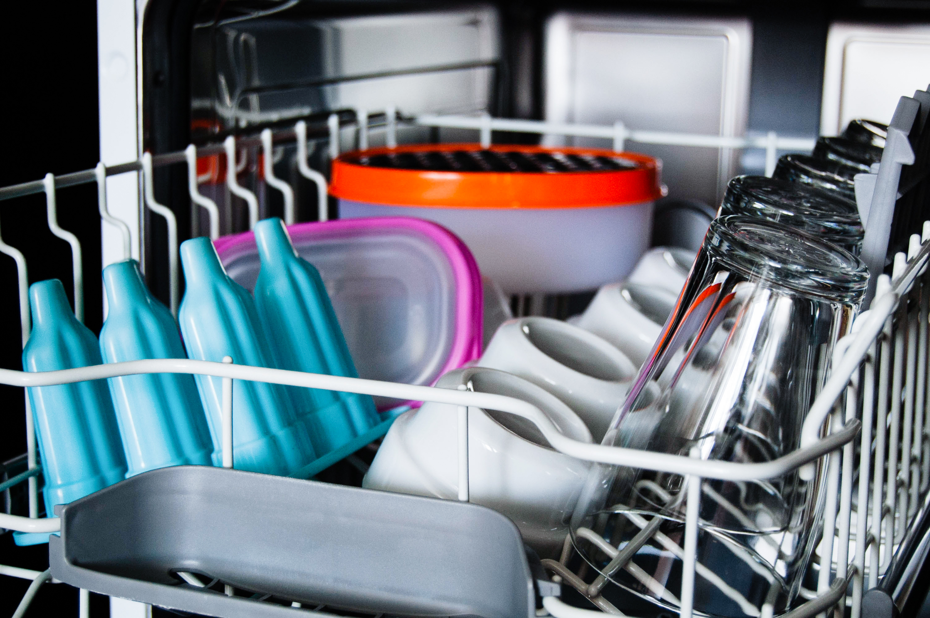 The top rack of a dishwasher filled with glasses, white cups, and colourful plastic containers.