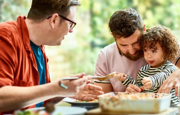 Three people sit at a table sharing a meal, with one adult guiding a child scoop food from a casserole dish while another adult holds a serving spoon.