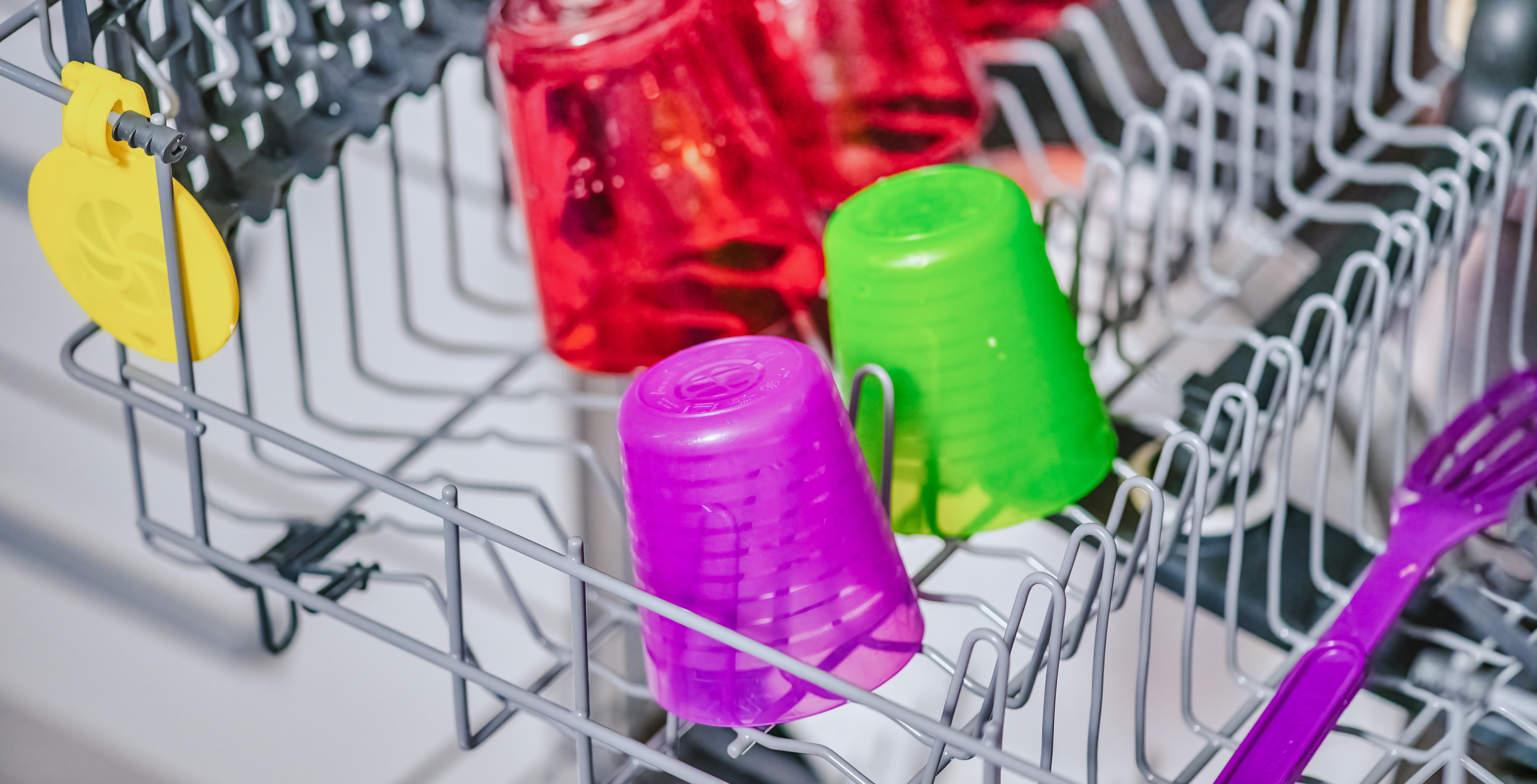 Des gobelets et des ustensiles en plastique de couleurs vives placés dans un panier de lave-vaisselle avant le nettoyage.