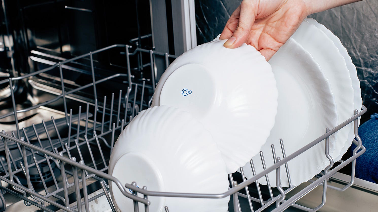 A woman’s hand removes a white bowl with a dishwasher symbol on it from a loaded dishwasher.
