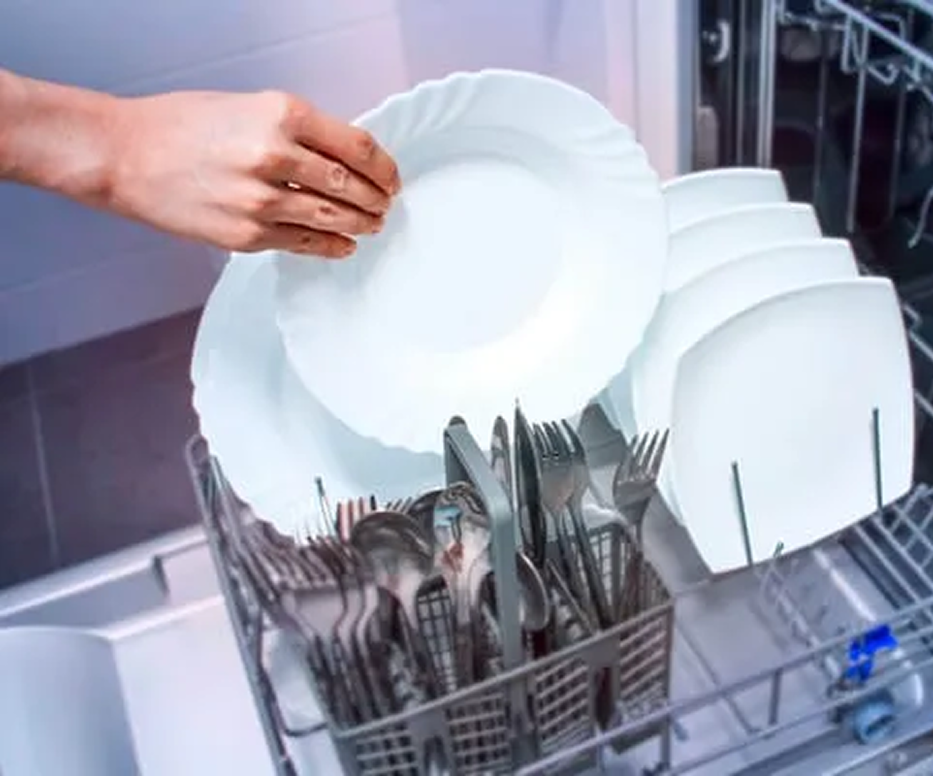 A hand takes out a clean, white plate from a dishwasher that is filled with neatly arranged plates and utensils.
