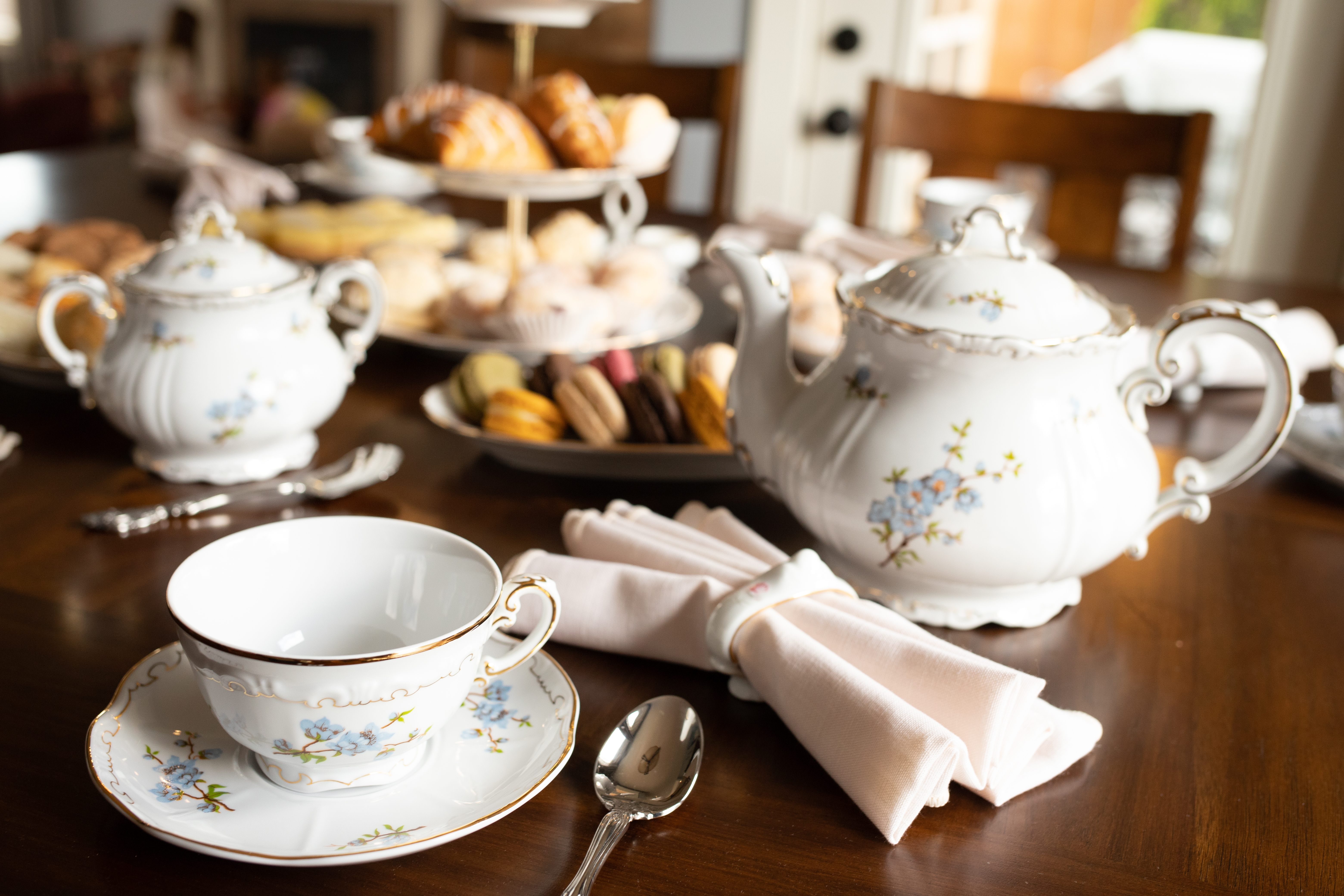 A table laid with a white teacup and saucer, teaspoon, napkin, teapot, sugar pot, and cookies.