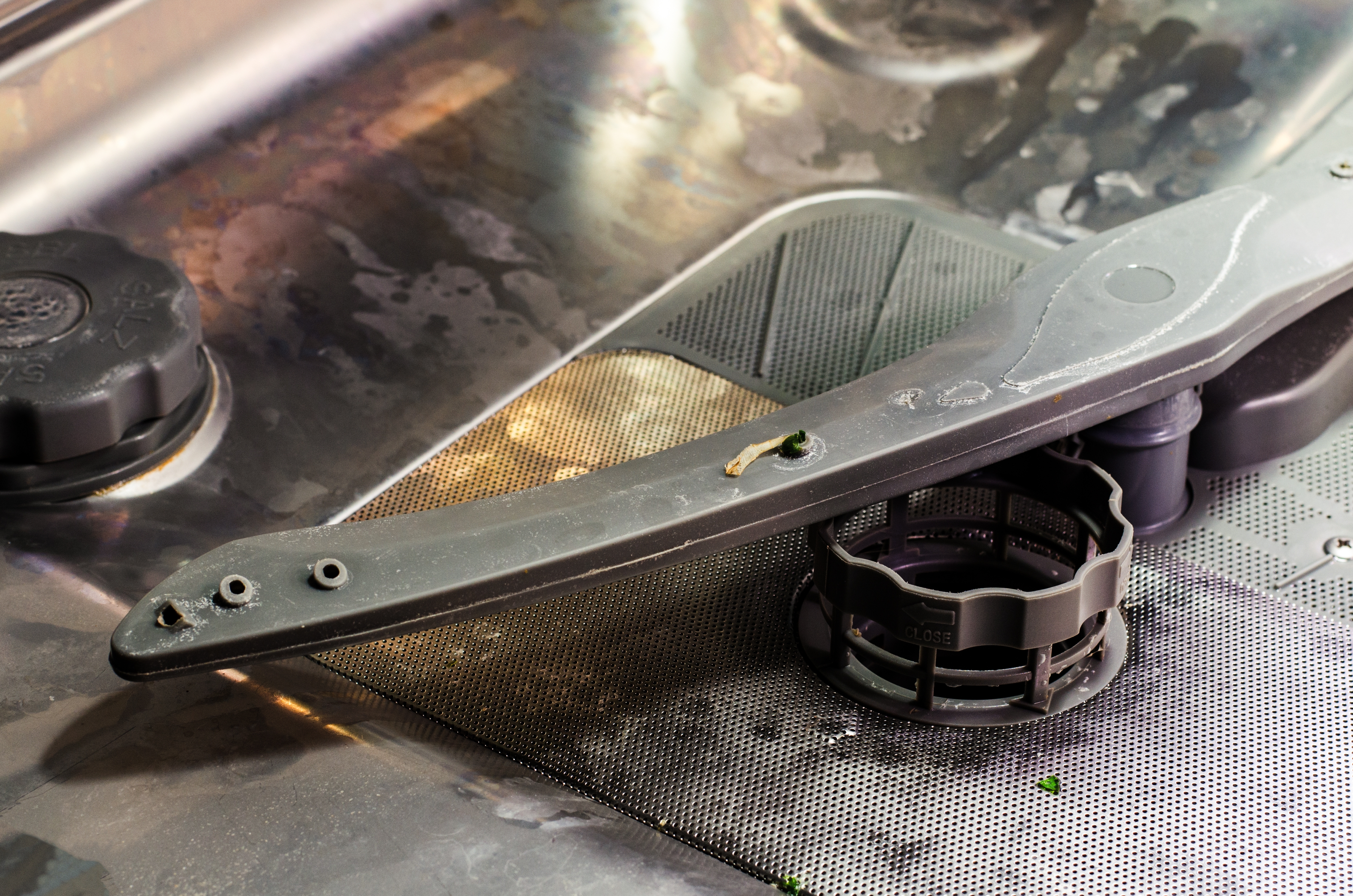 Close-up of a dishwasher's interior, showing the wet metallic surface, spray arm, and filter with food debris.