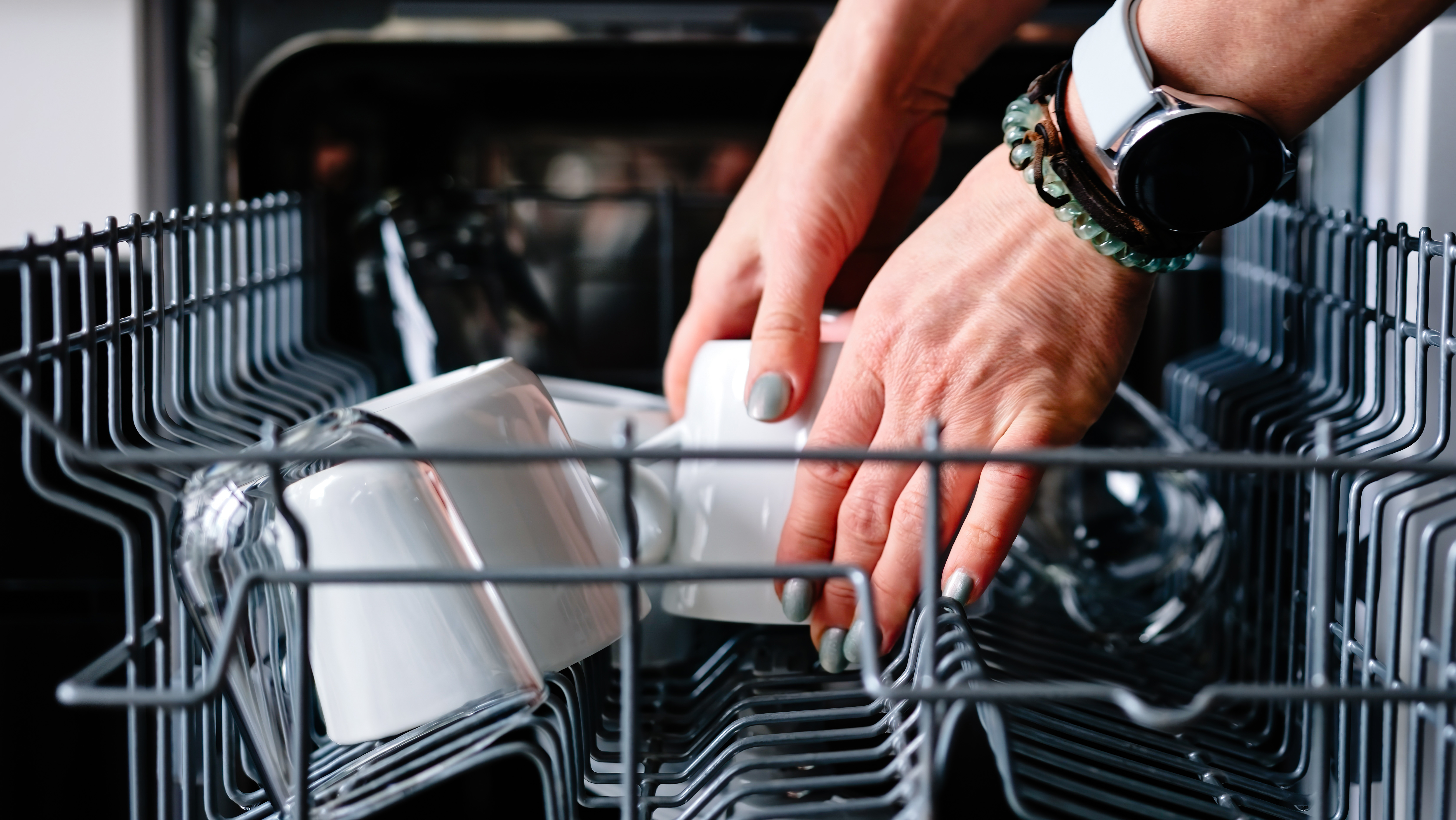 A woman’s hands take a white cup out of the top rack of a dishwasher.