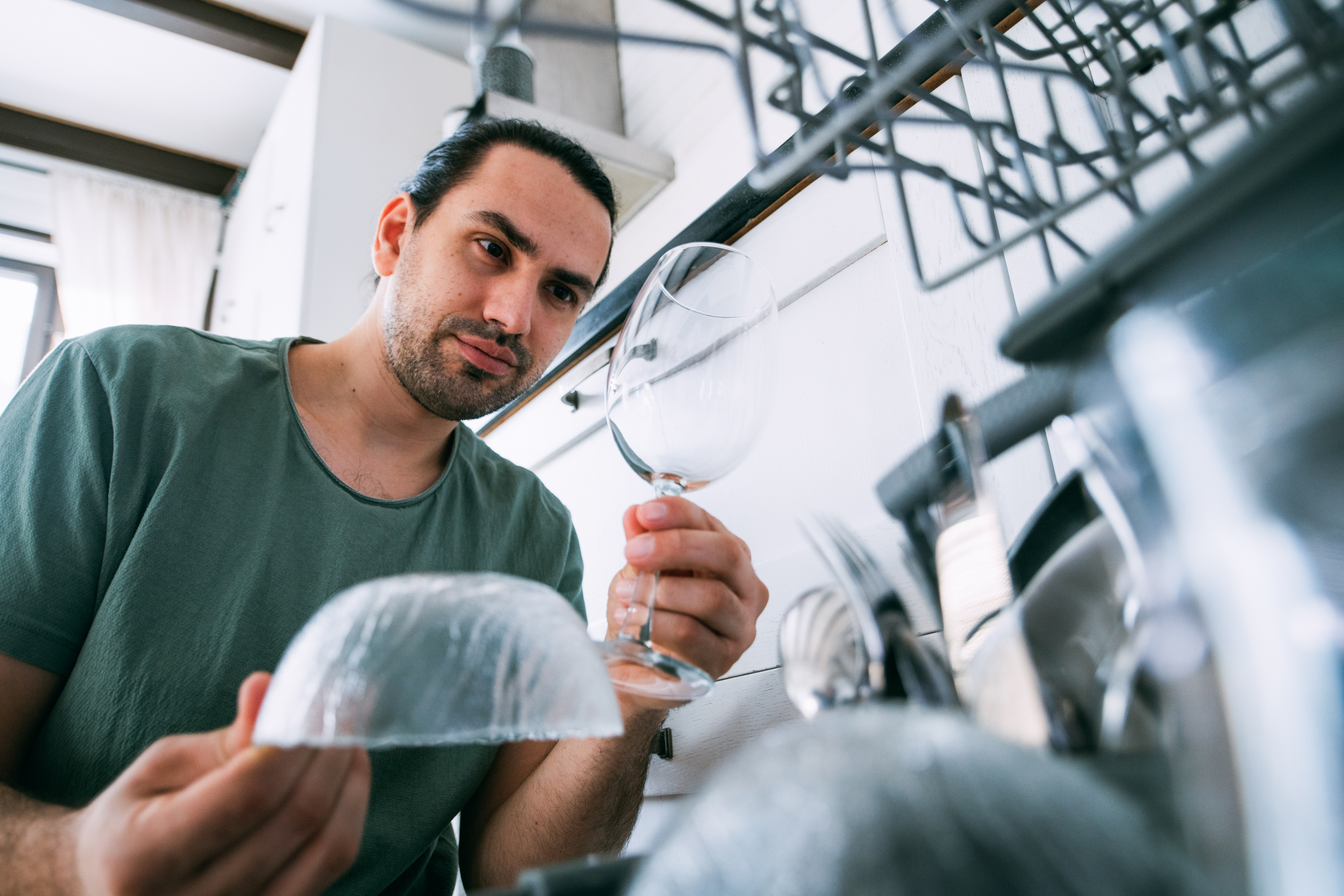 Homme inspecte un verre de vin propre et un bol pendant qu’il décharge un lave-vaisselle dans une cuisine moderne.