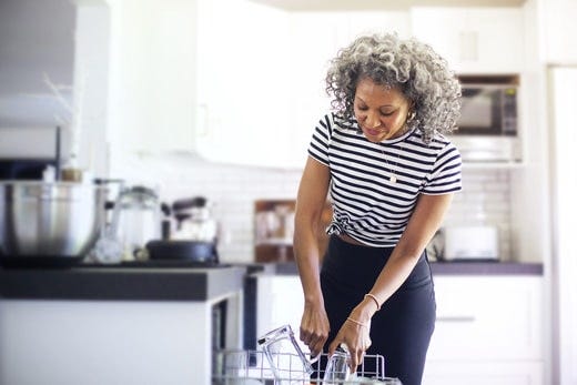 A woman in a striped t-shirt loads glasses into the top rack of a dishwasher in a white kitchen.