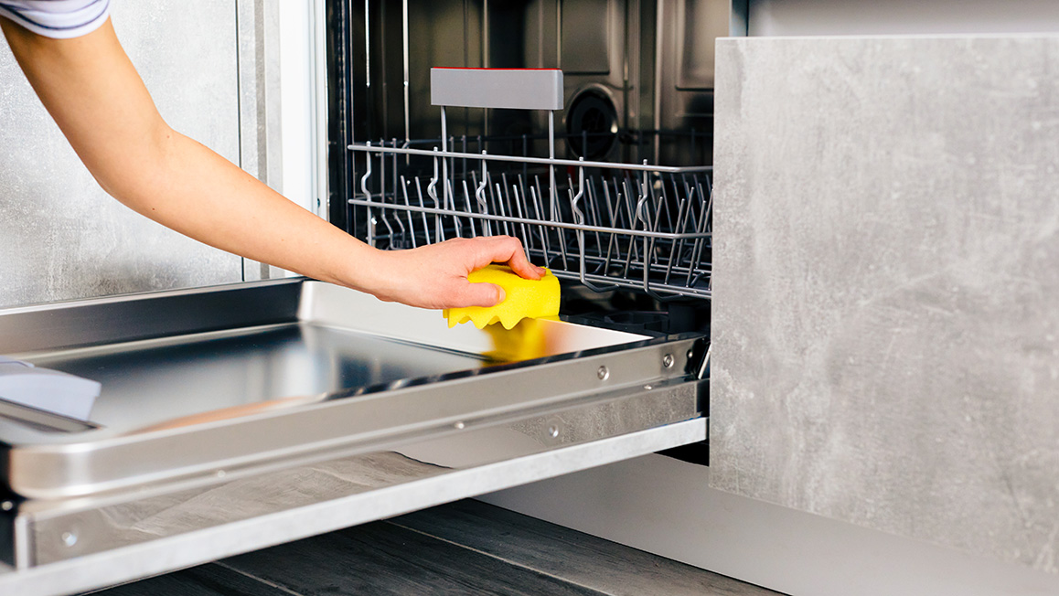 A woman’s hand holds a yellow sponge on the inside of a dishwasher door that is laid flat.