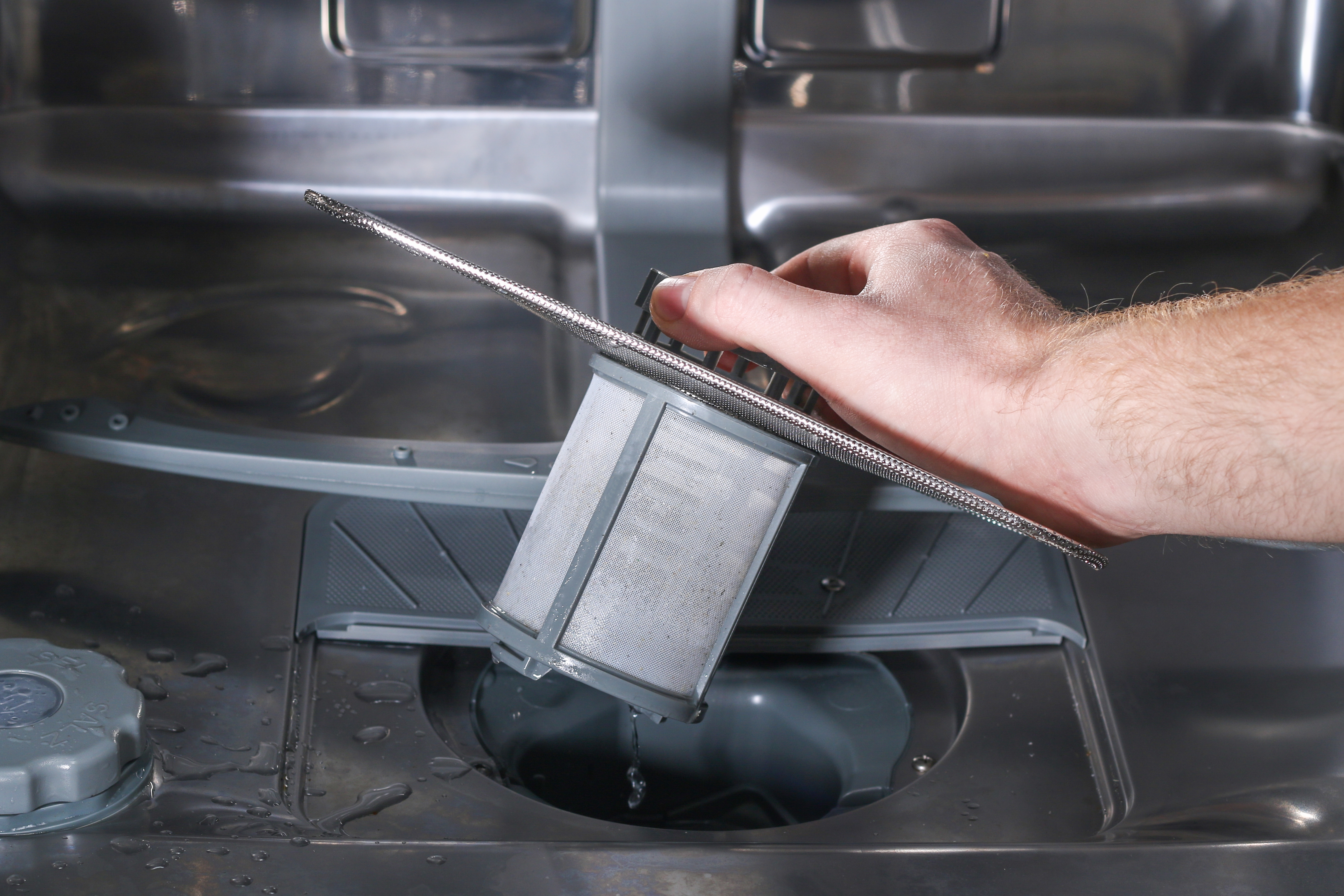 A hand removing the circular mesh dishwasher filter from the base of the machine.