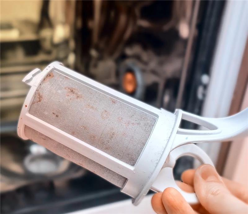 A hand holding a dishwasher filter removed from the dishwasher with slight grime visible.