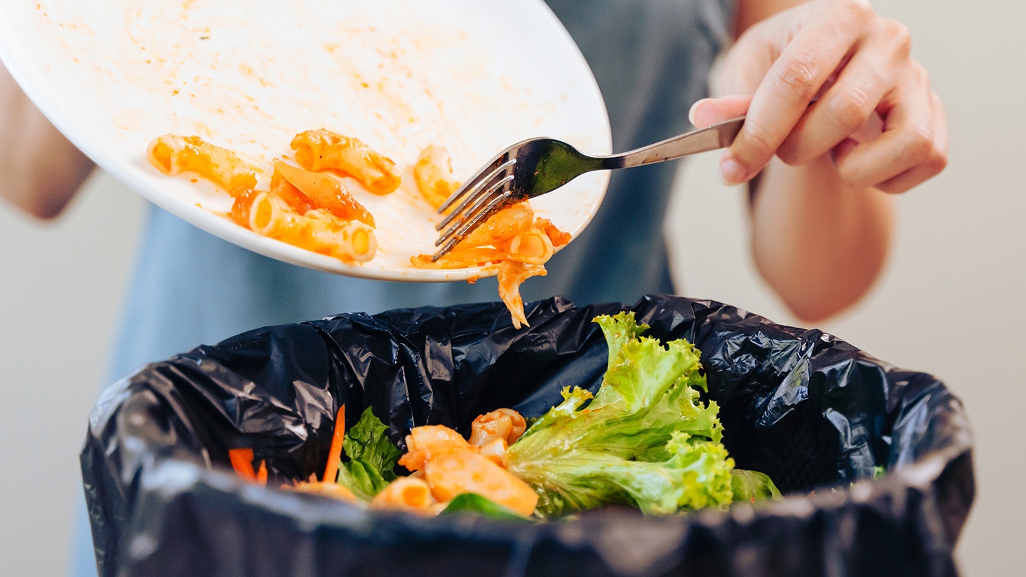 A woman’s hand scrapes leftover pasta from a white dish into a garbage container filled with discarded vegetables.