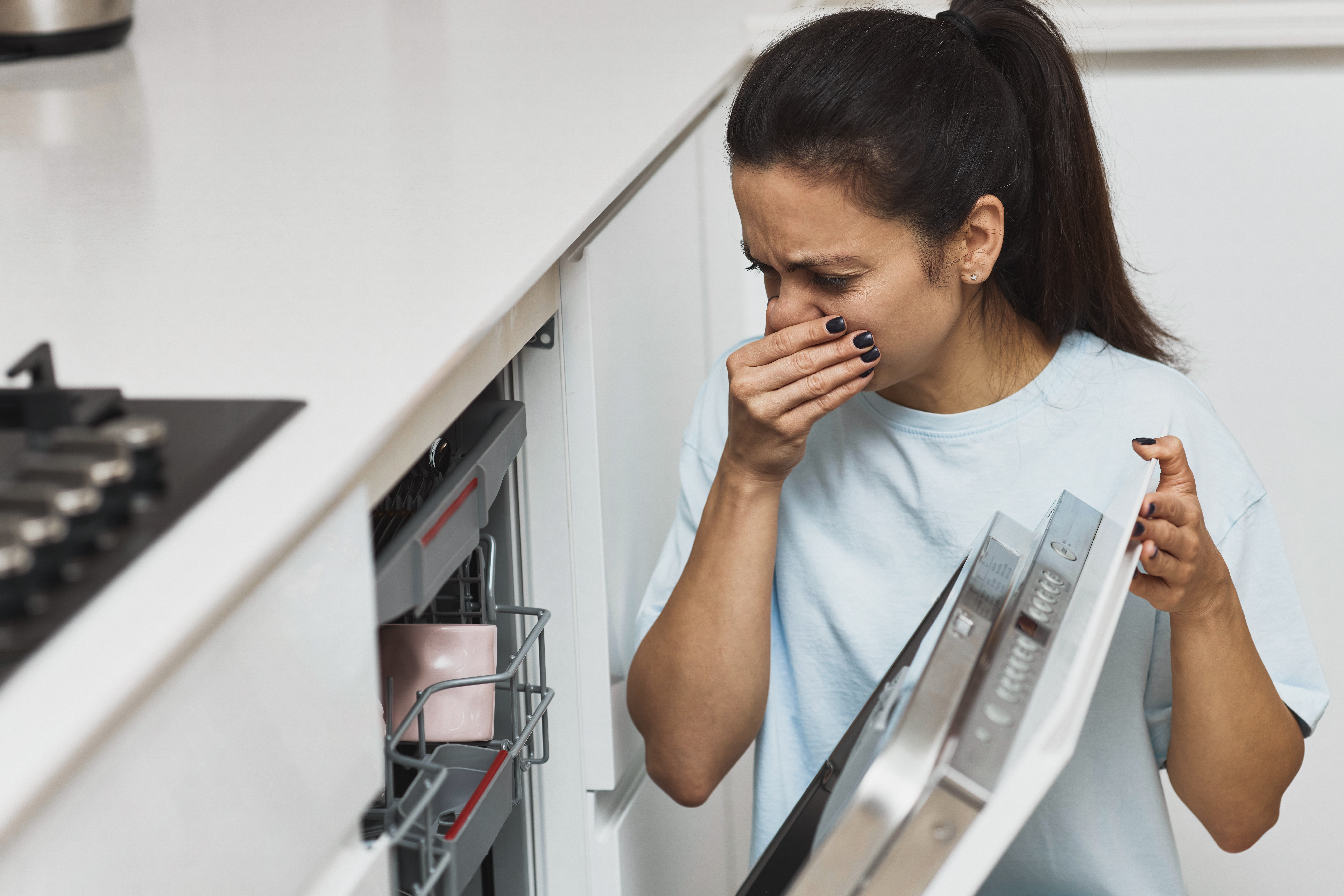 A woman wearing a white shirt holds a dishwasher door open with one hand while covering her nose with the other, reacting to an unpleasant smell.