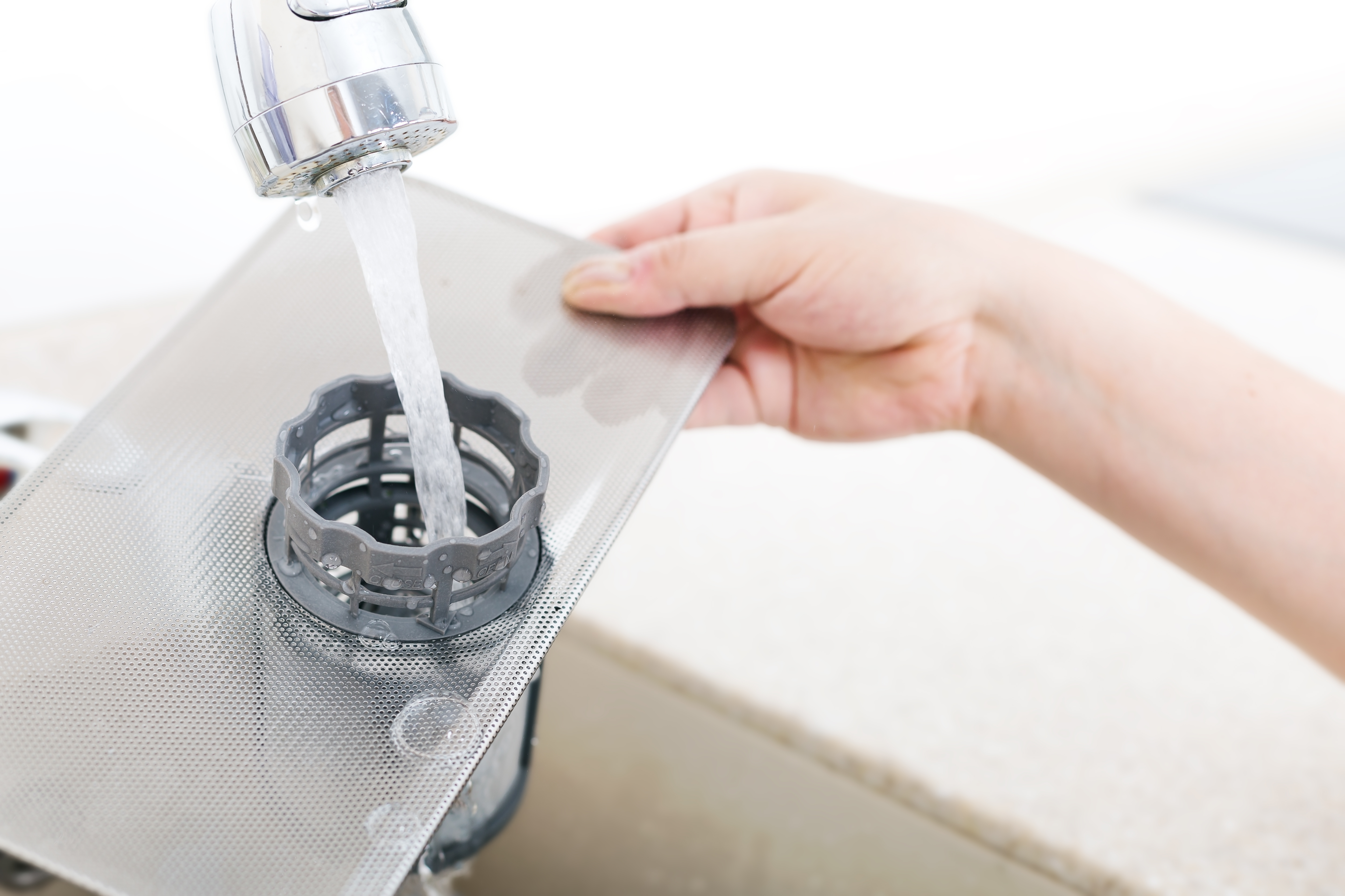 A hand holding a removed dishwasher filter and washing it under running water.