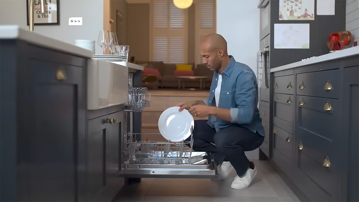 A man squats on the ground, removing a white dish from a dishwasher in a grey kitchen.