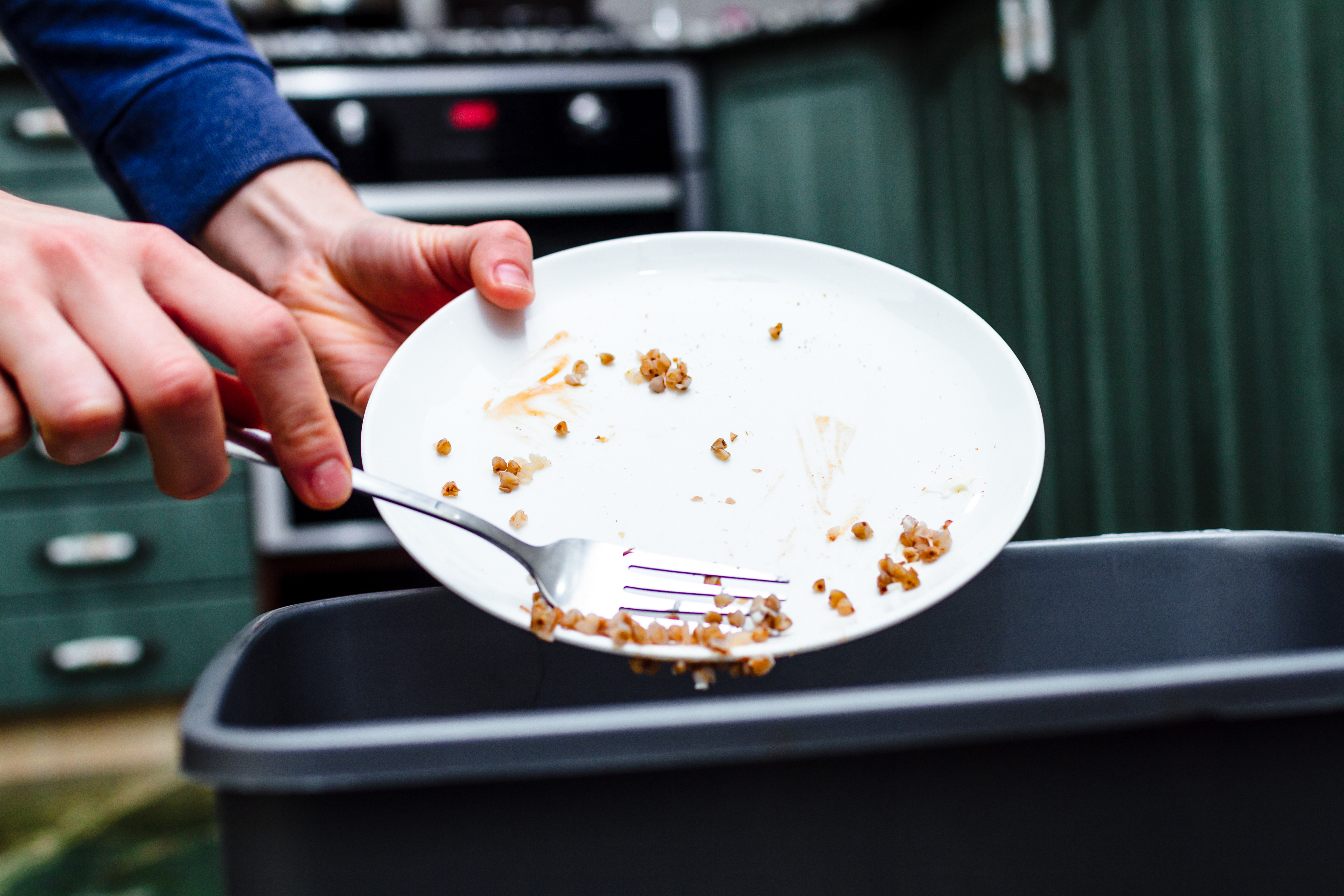 One hand holds a white plate while the other hand uses a fork to scrape food into a garbage container.