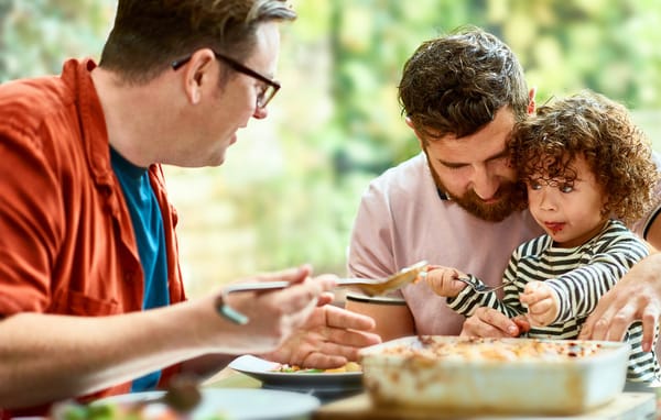 Trois personnes sont assises à une table partageant un repas, un adulte guidant un enfant qui prend de la nourriture dans un plat à gratin tandis qu'un autre adulte tient une cuillère de service.