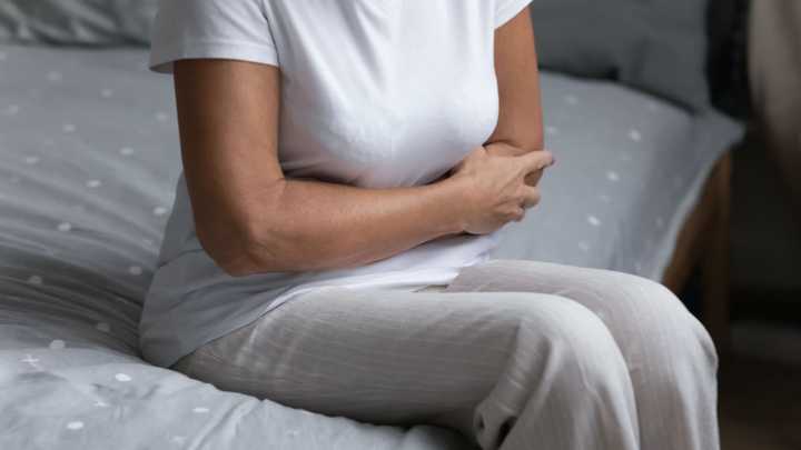 Person sitting on the edge of a bed, leaning forward and holding their abdomen as if experiencing stomach pain